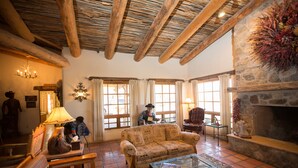 A cozy living room with a stone fireplace, wooden beams, and a large window with curtains. - Tanque Verde Ranch (Tucson)