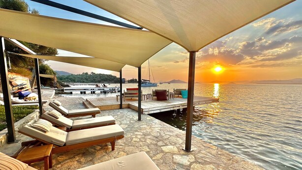 A sunlit outdoor seating area with lounge chairs and a wooden deck overlooking a body of water. - Ece Hotel Sovalye Island (Fethiye)