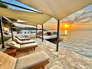 Un espacio al aire libre con sol, sillas reclinables y una terraza de madera con vista a un cuerpo de agua.