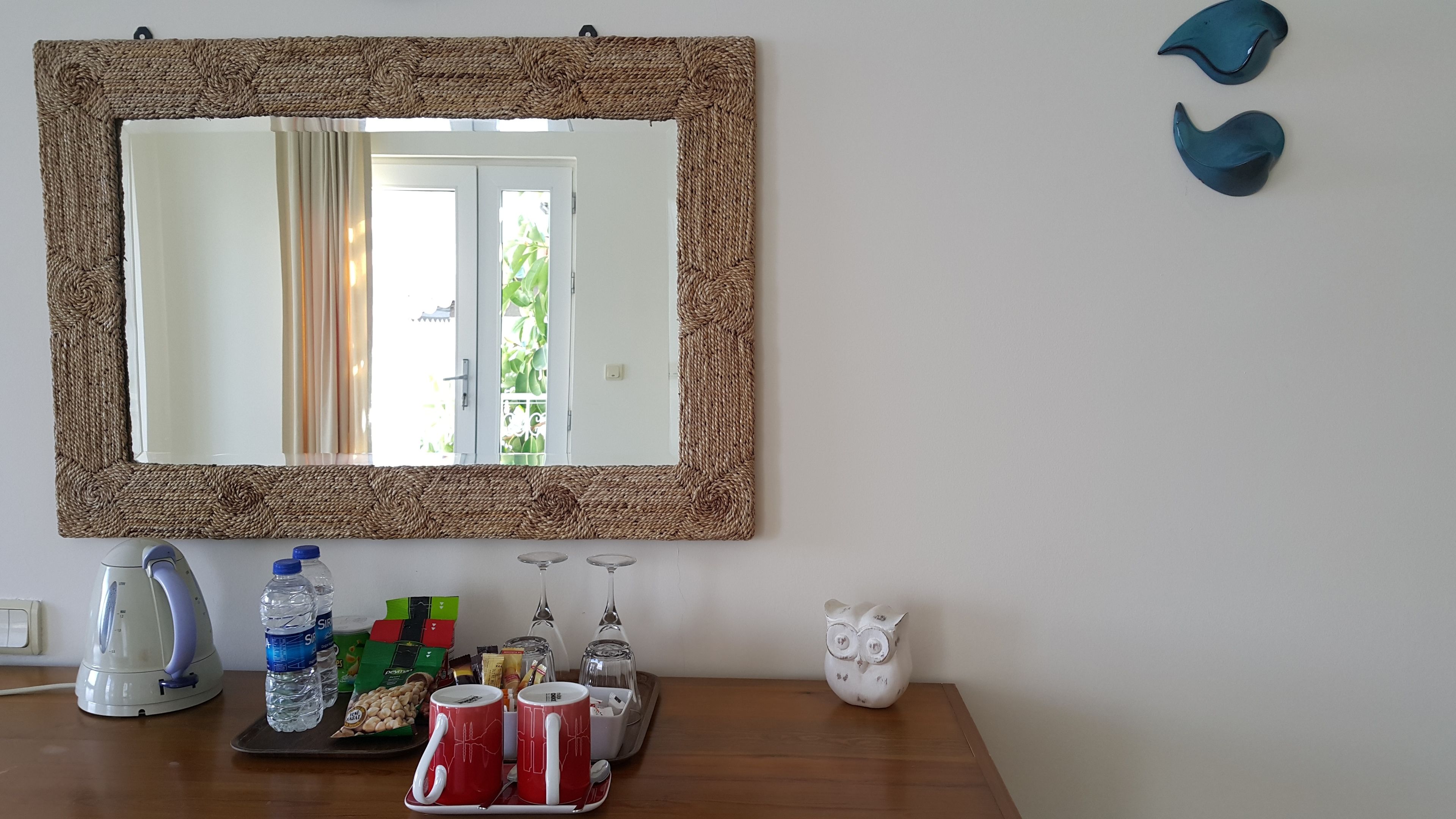 A mirror with a woven frame, a kettle, water bottles, a tray with snacks, and a white owl figurine on a wooden table.