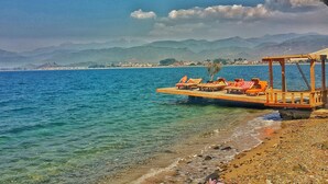 A wooden pier with lounge chairs extending over clear blue water, a pebble beach, and a distant mountain range. - Ece Hotel Sovalye Island (Fethiye)