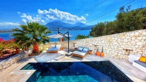 A poolside area with a stone wall, lounge chairs, and a view of the sea and mountains. - Ece Hotel Sovalye Island (Fethiye)