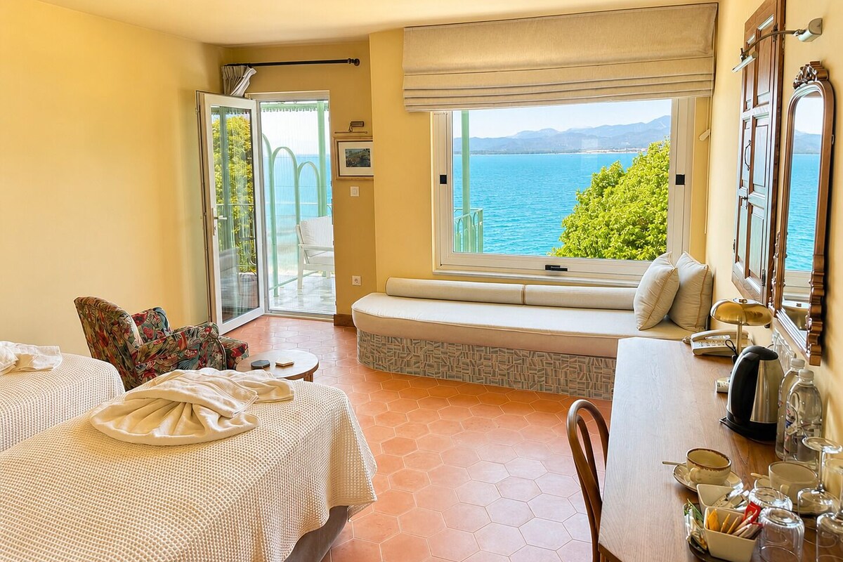 A room with two beds, a dining table, a window with a view of the sea, and a balcony with a chair.