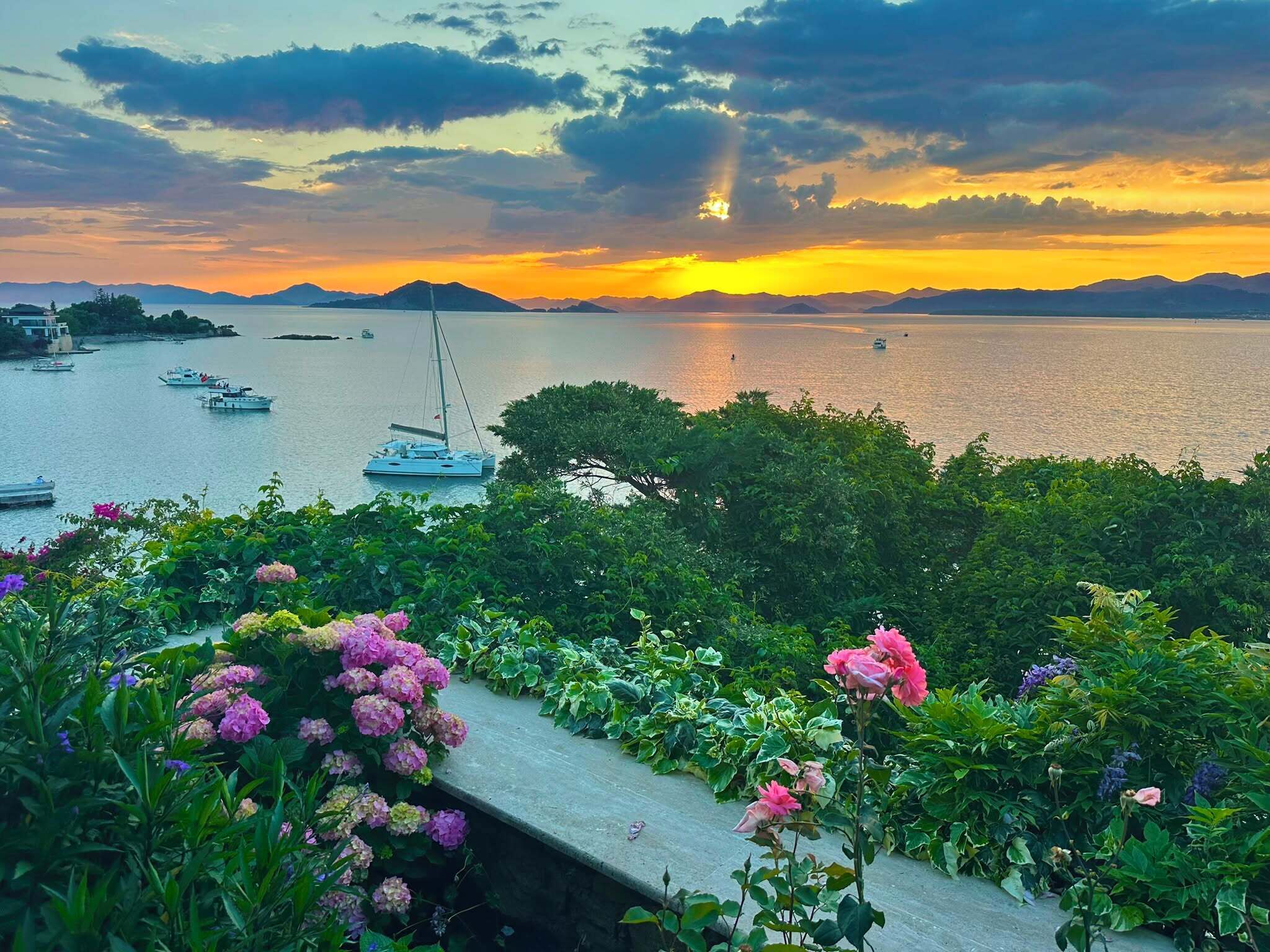 Vista costera con barcos, un velero y un jardín lleno de flores.