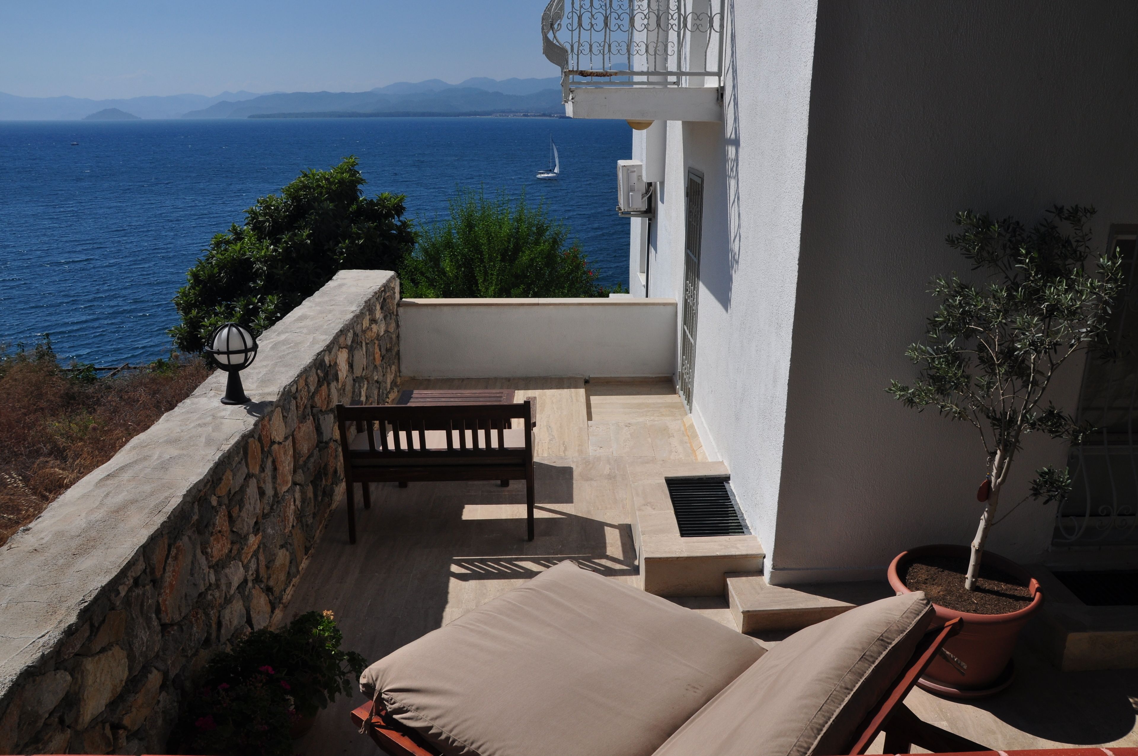 A balcony with a stone wall, a bench, a potted plant, and a view of the sea.