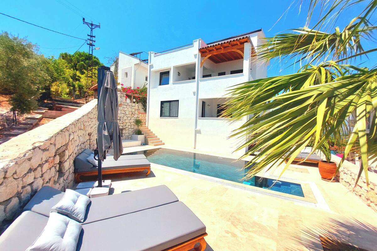A modern villa with a swimming pool, stone walls, and a balcony with lounge chairs.