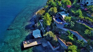 An aerial view of a coastal property with a stone wall, lush greenery, and a wooden dock extending into clear blue waters. - Ece Hotel Sovalye Island (Fethiye)