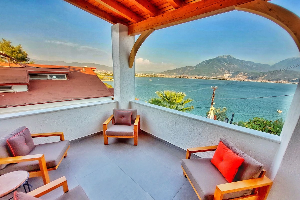 A balcony with wooden furniture, a view of a body of water, and mountains in the distance.