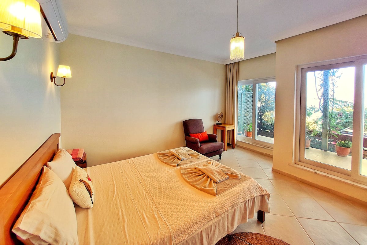 A hotel room with a bed, a chair, a small table, and a large window with a view of the outdoors.