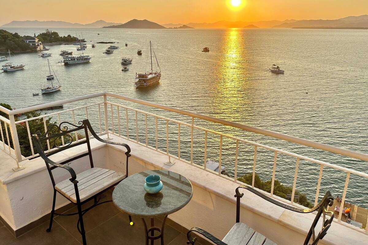 A balcony with a table and chairs overlooking a marina with boats and a sunset.
