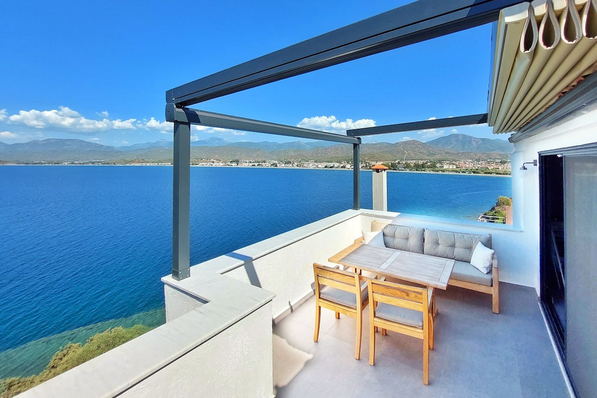 A balcony with a view of the sea and mountains, a wooden table, and a cushioned seating area.