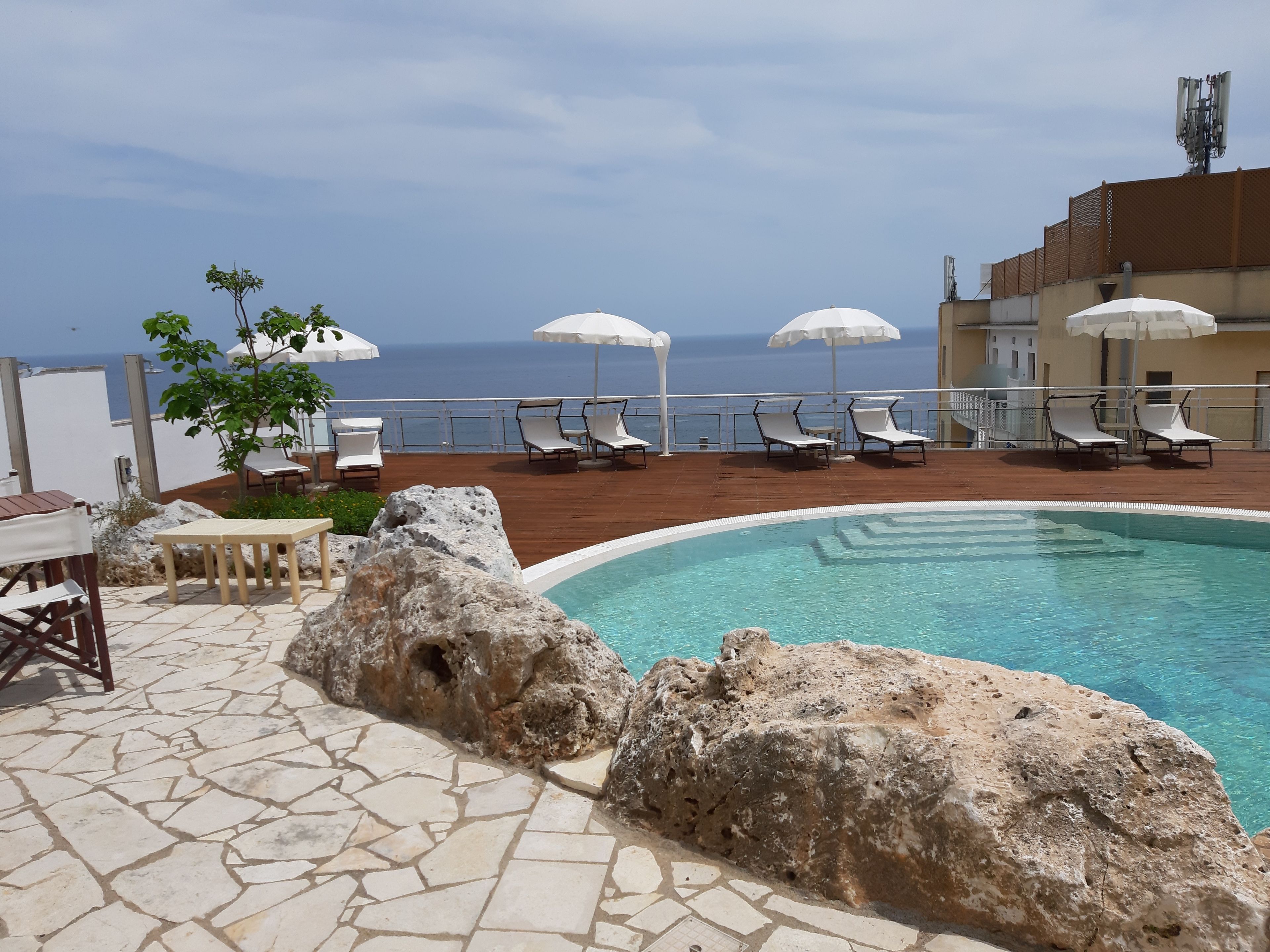 Un área de piscina con sillones de descanso y sombrillas, suelo de piedra y vista al mar.