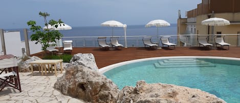 Un área de piscina con sillones de descanso y sombrillas, suelo de piedra y vista al mar.