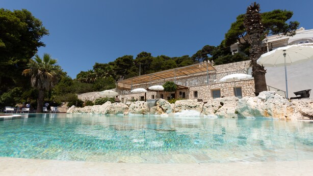 Piscina cristalina con un edificio de piedra al fondo, rodeada de árboles y rocas.