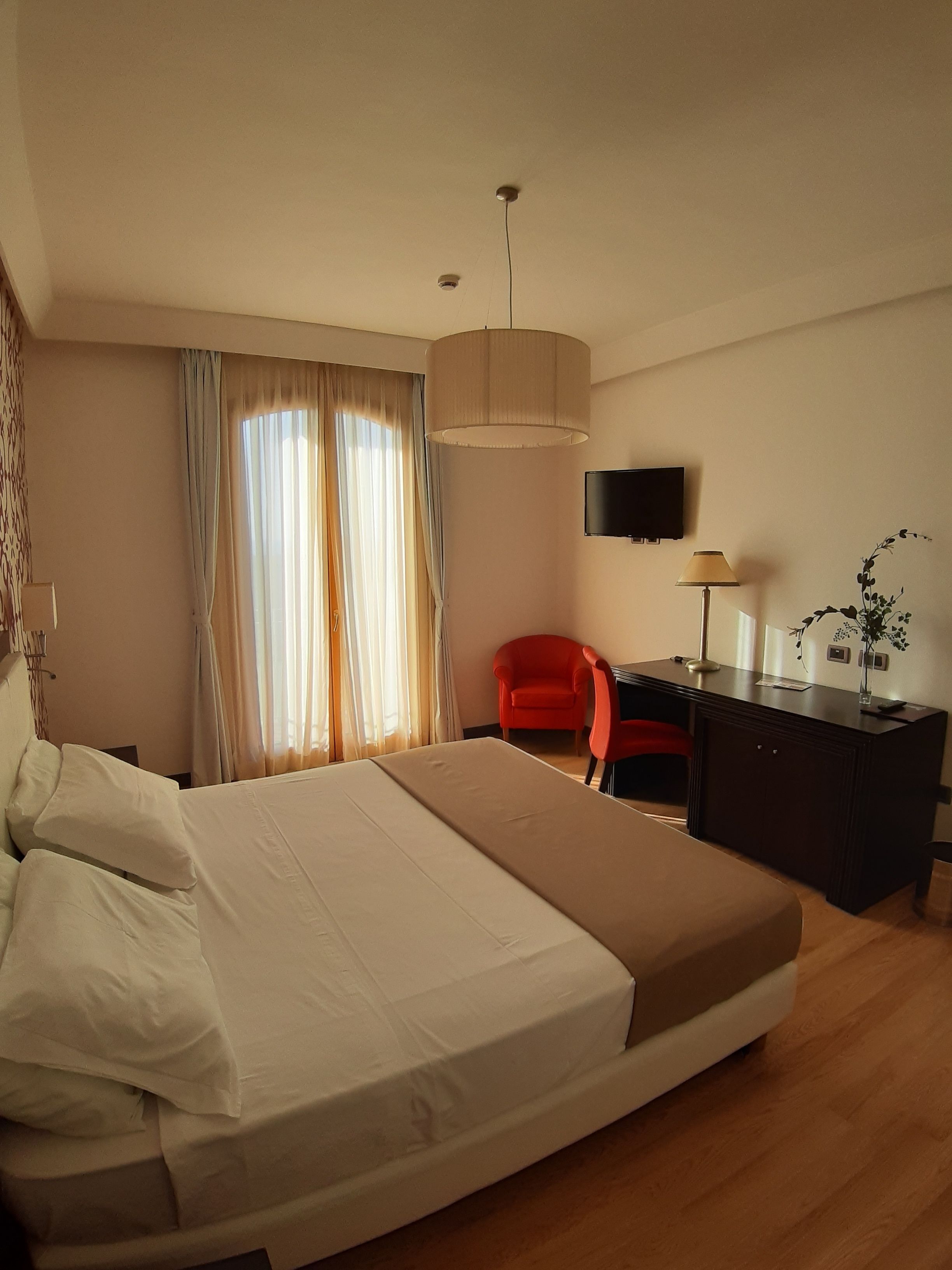Quarto de hotel com uma cama, escrivaninha, cadeira, televisão e janela com cortinas.