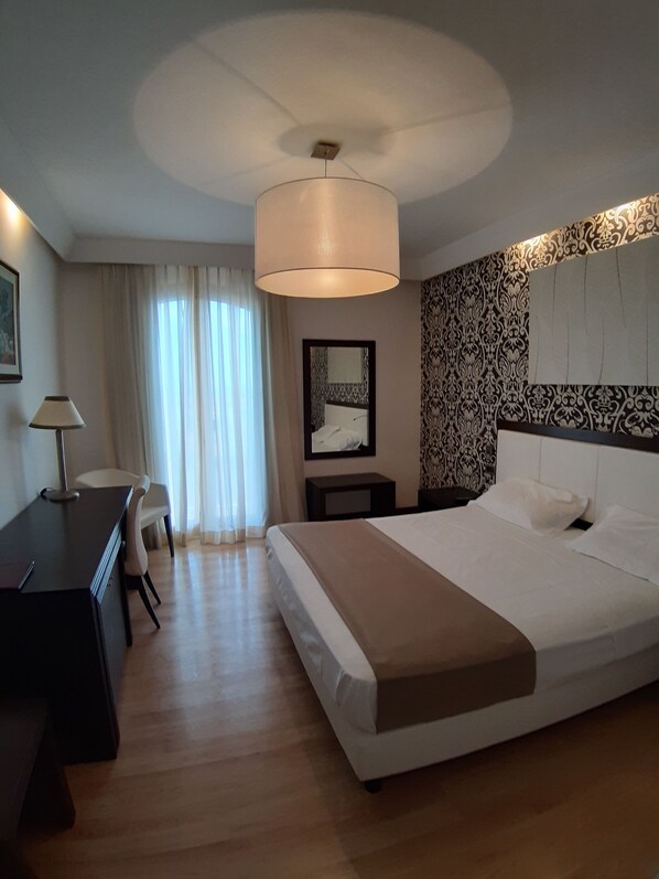 Une chambre d’hôtel avec un lit, un bureau, une chaise, une lampe et une grande fenêtre avec des rideaux.