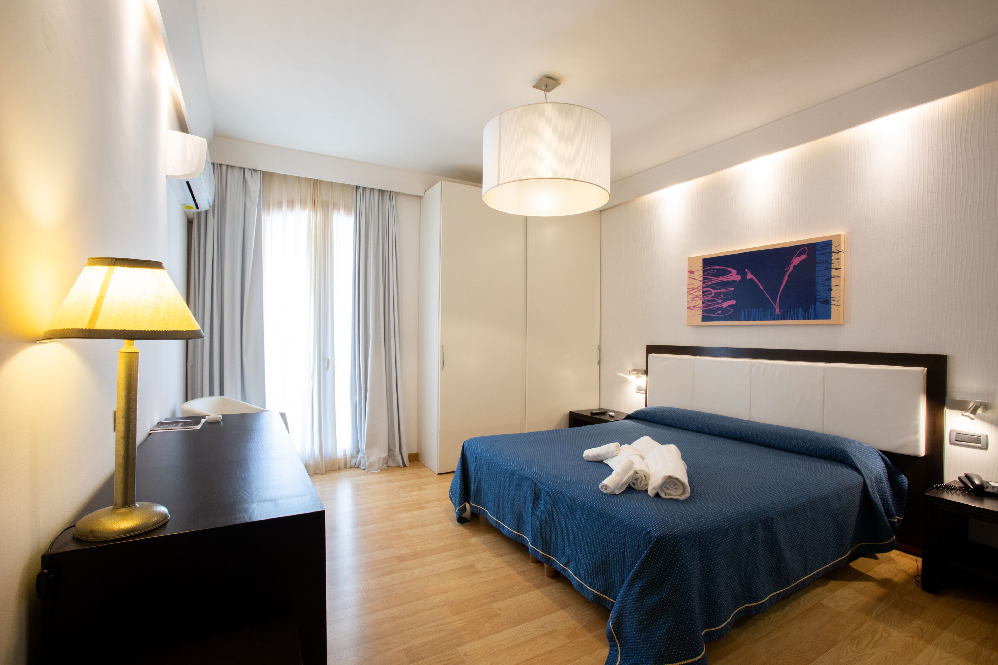 A hotel room with a bed, bedside table, lamp, and a painting on the wall.