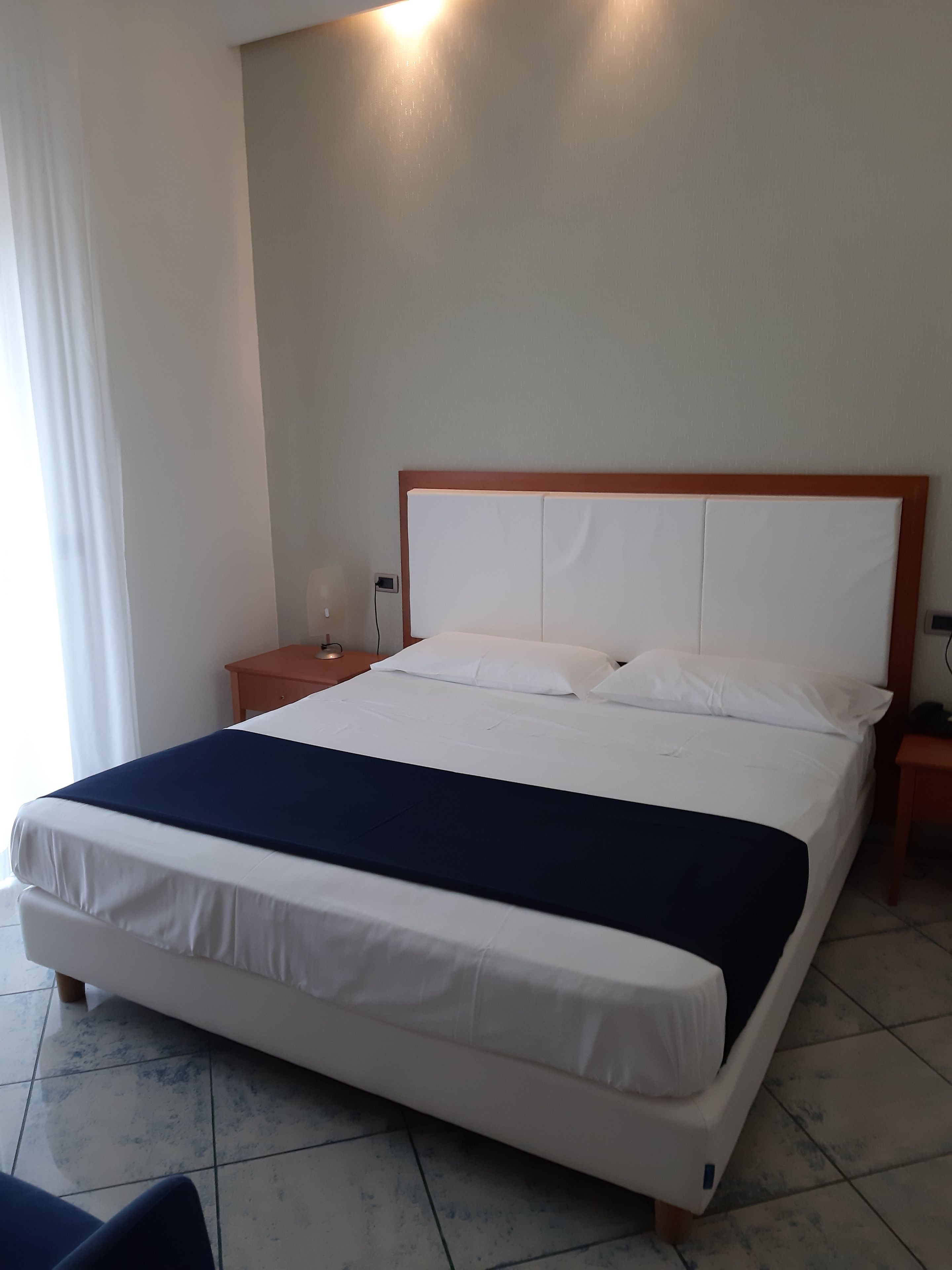 A neatly made bed with white linens and a dark blue blanket, a wooden nightstand with a lamp, and a wall-mounted light fixture.