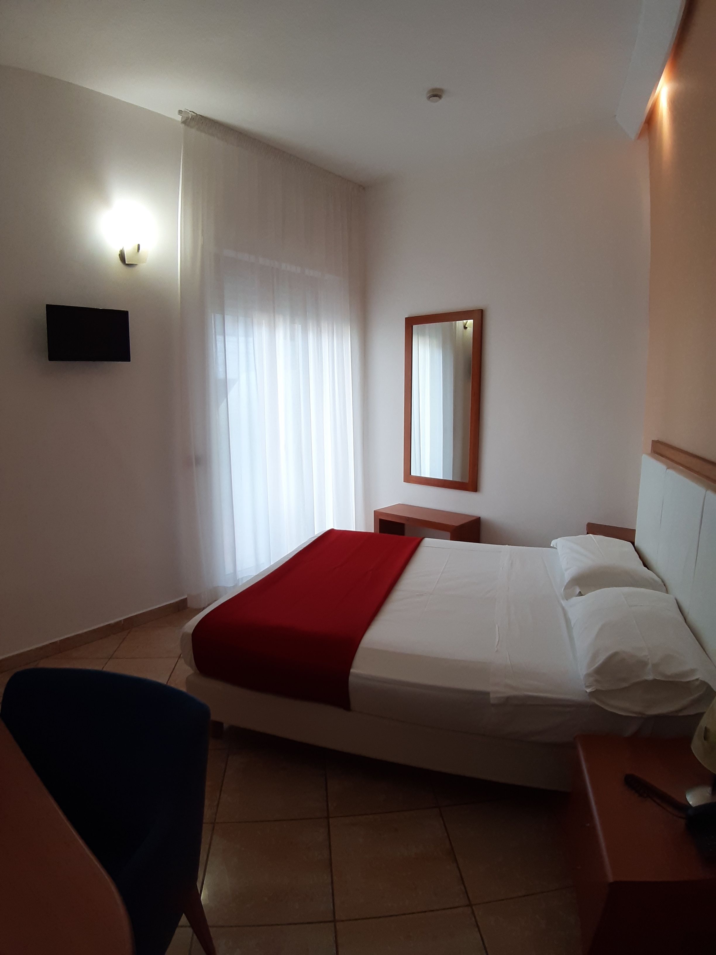 Ein Hotelzimmer mit Bett, Sessel, Nachttisch, Spiegel und einem an der Wand befestigten Fernseher.