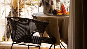 Un balcón con una silla de mimbre, una mesita redonda con mantel, una jarrón y un plato de frutas.