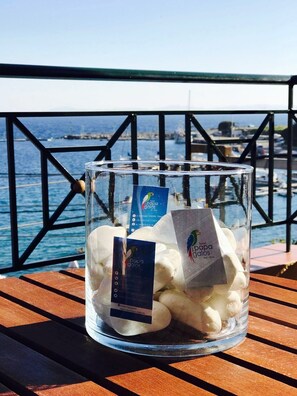 A glass jar filled with white stones on a wooden bench with a metal railing and a view of the sea and boats in the background. - Hotel Papagalos (Sithonia)