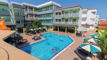 A swimming pool surrounded by multi-story buildings with balconies, sun loungers, and umbrellas.