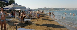 Ein Strand mit Liegestühlen und Sonnenschirmen, Menschen beim Schwimmen und Spazierengehen am Ufer.