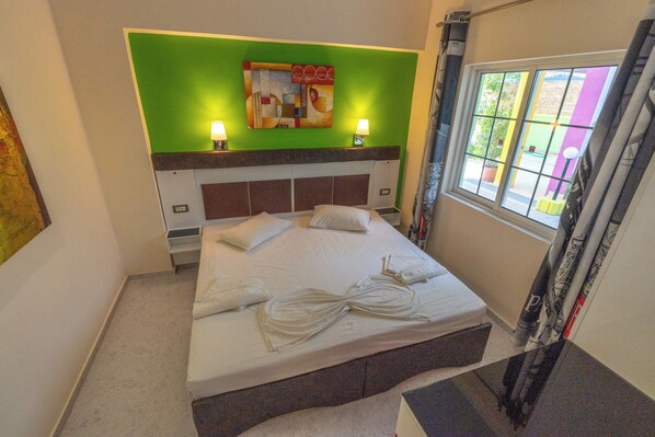 Quarto de hotel com cama, almofadas, parede verde de destaque e janela com cortinas.