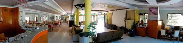 A spacious hotel lobby with a central seating area, multiple dining tables, and a large window providing natural light.