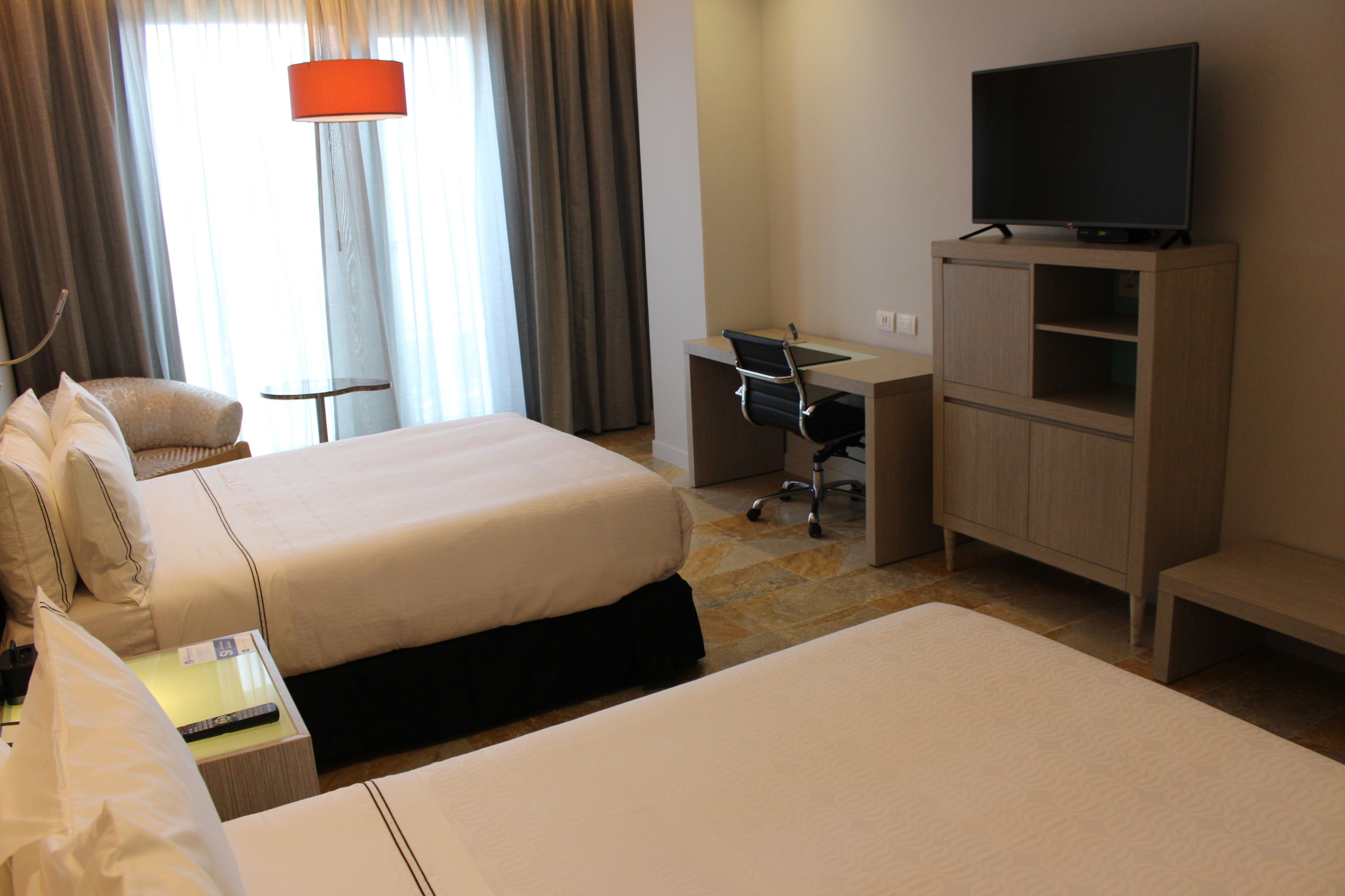 A hotel room with two beds, a desk, a chair, a television, and a lamp.