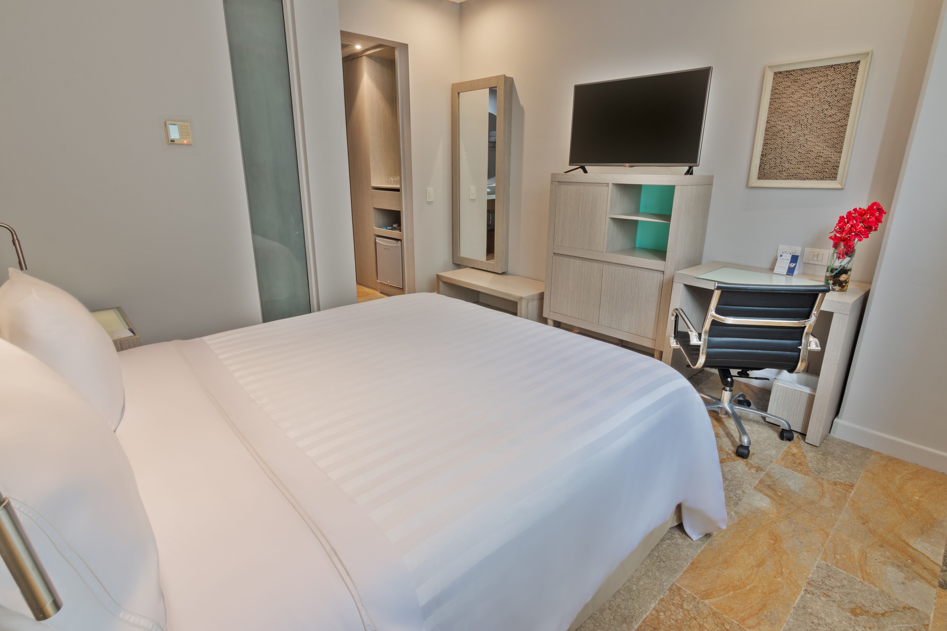 A hotel room with a large bed, a desk with a chair, a TV, a mirror, and a small bathroom area.