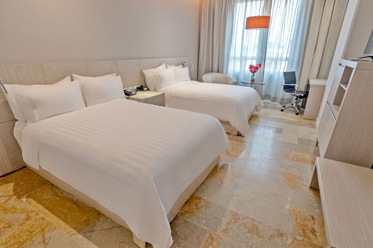 A hotel room with two beds, a desk, a chair, and a lamp.