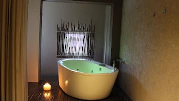 A bathtub in a room with a wooden floor, a candle, and a metal fence in the background.