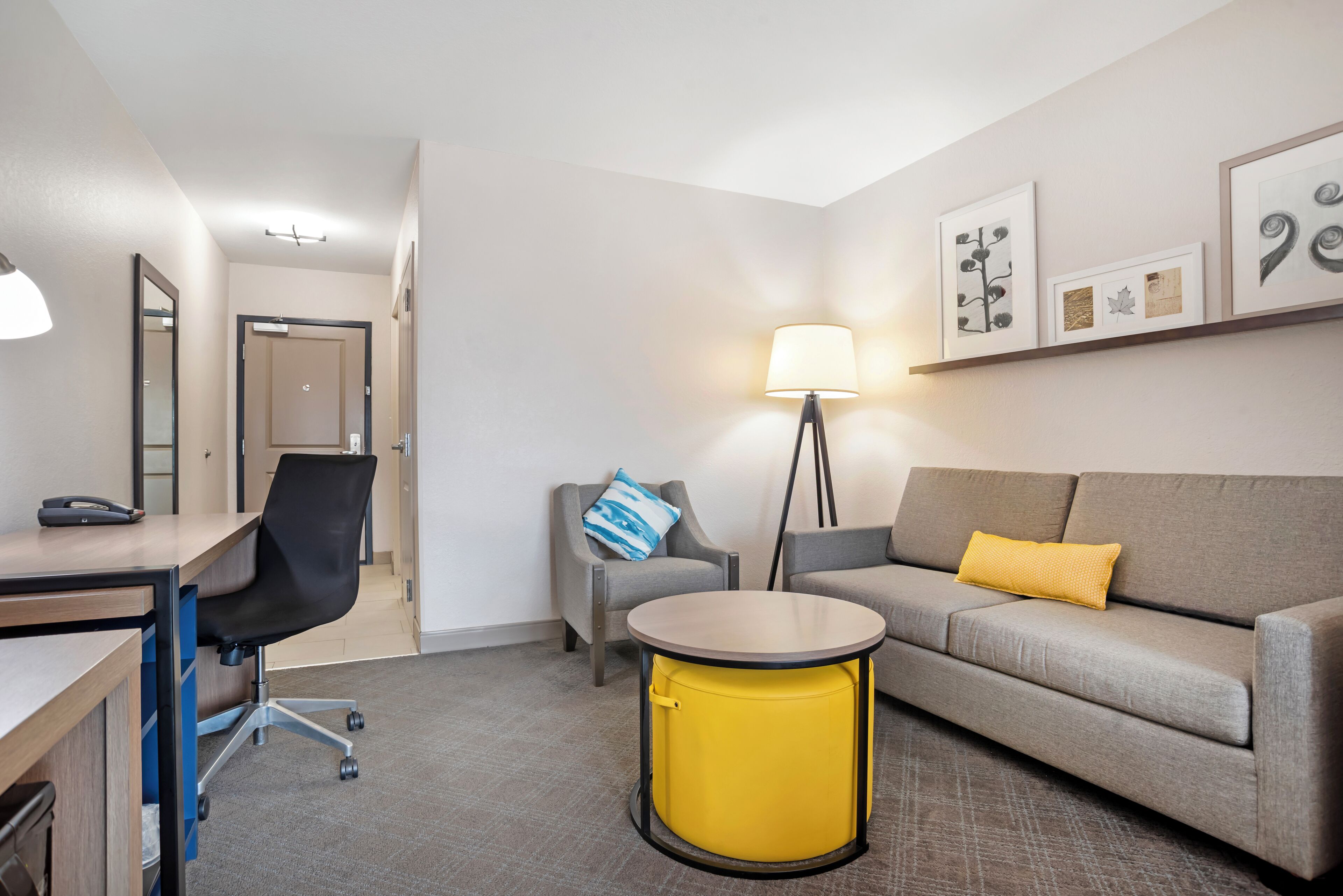 A modern hotel room with a desk, office chair, round yellow table, grey sofa, and a lamp.