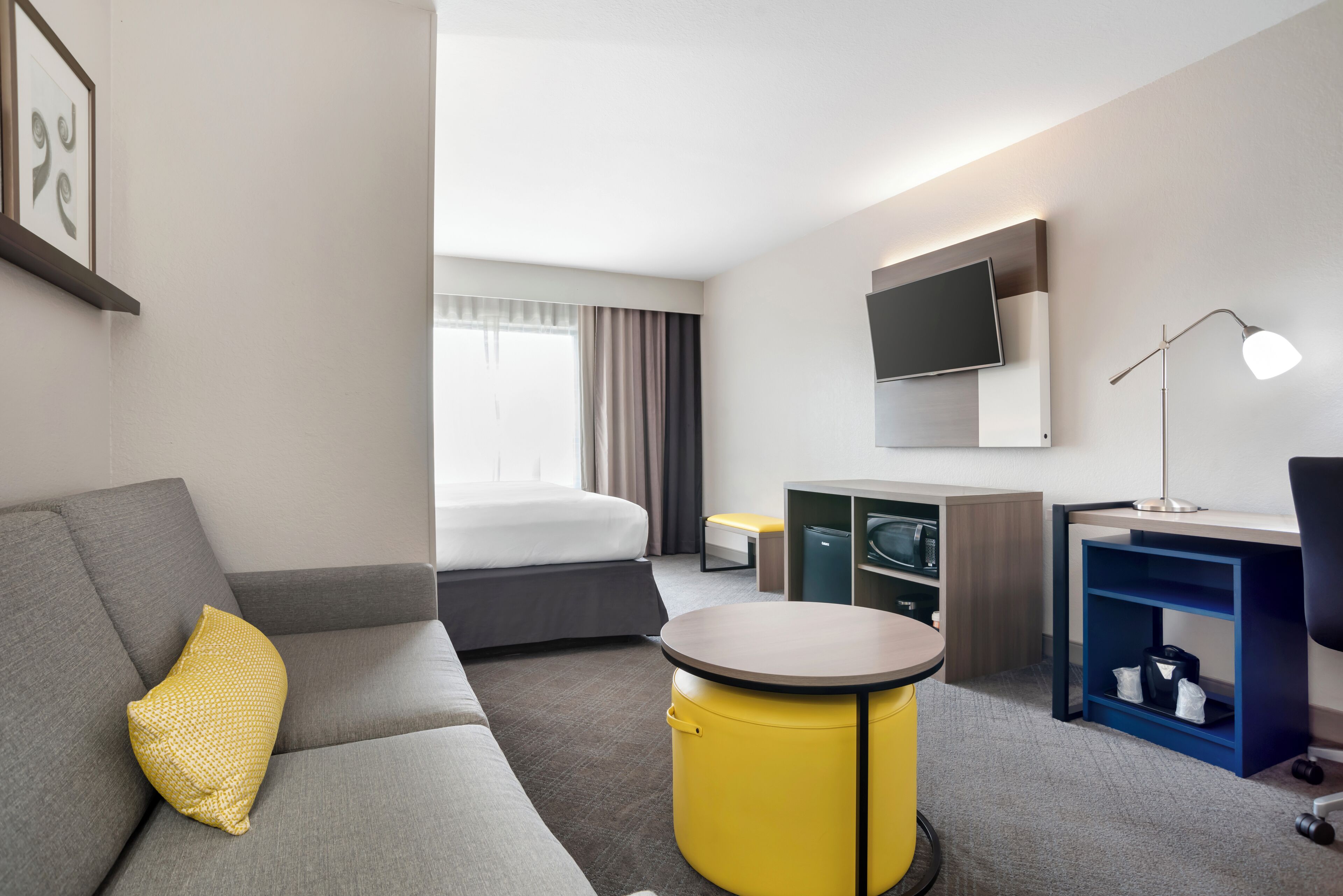 A modern hotel room with a grey sofa, a yellow ottoman, a bed with grey bedding, a flat-screen TV mounted above the bed, a desk with a lamp, and a blue shelving unit.