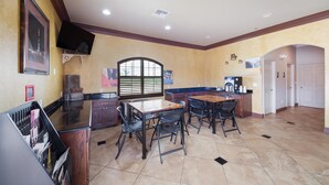 A spacious room with a large dining table, chairs, a TV mounted on the wall, and a window with blinds. - Eagle Ford Inn (Floresville)