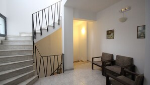 Un escalier avec des rampes noires menant à un palier, un coin salon avec deux fauteuils et un luminaire fixé au mur.