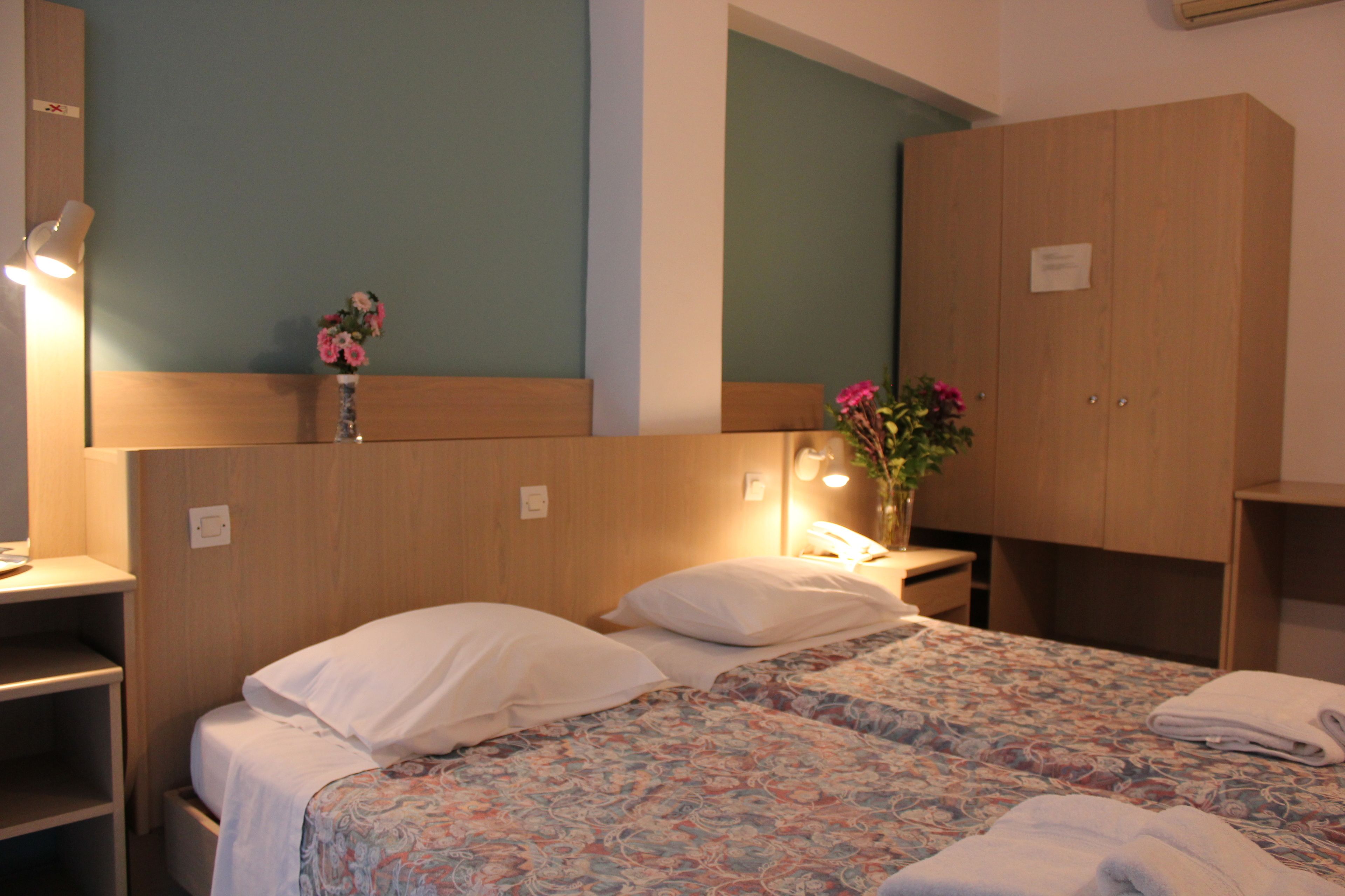 Une chambre d’hôtel avec deux lits, une armoire en bois, une table de chevet avec une lampe et un vase de fleurs.