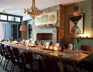 Un comedor con una larga mesa de madera dispuesta para una comida, una lámpara decorativa, una chimenea y una obra de arte grande enmarcada en la pared.