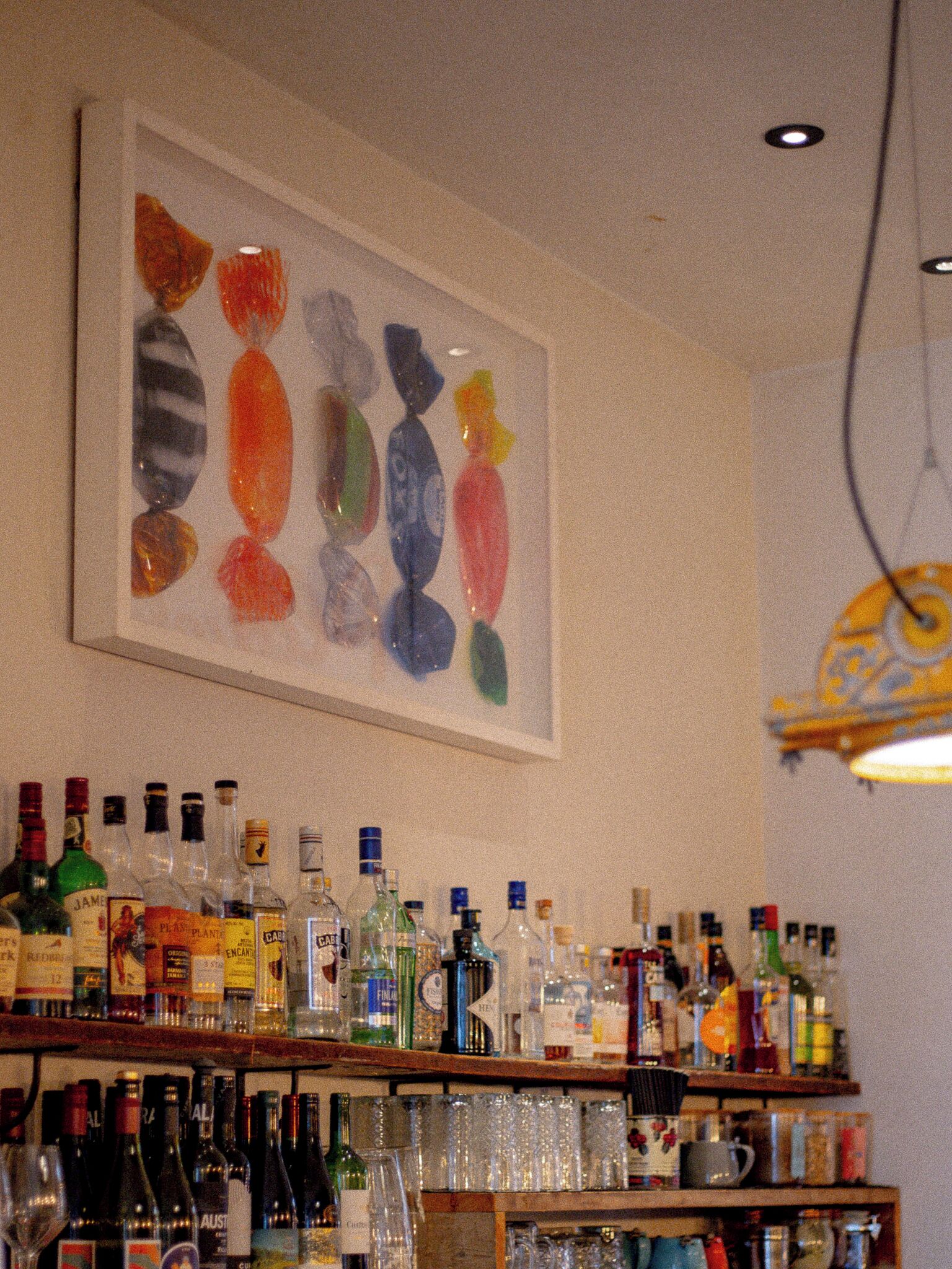 A bar area with various bottles and glasses on shelves, a painting of colorful shapes on the wall, and a hanging light fixture.
