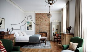 A bedroom with a bed, green armchairs, a wooden dresser, a brick wall, and a chandelier. - Artist Residence London (London)