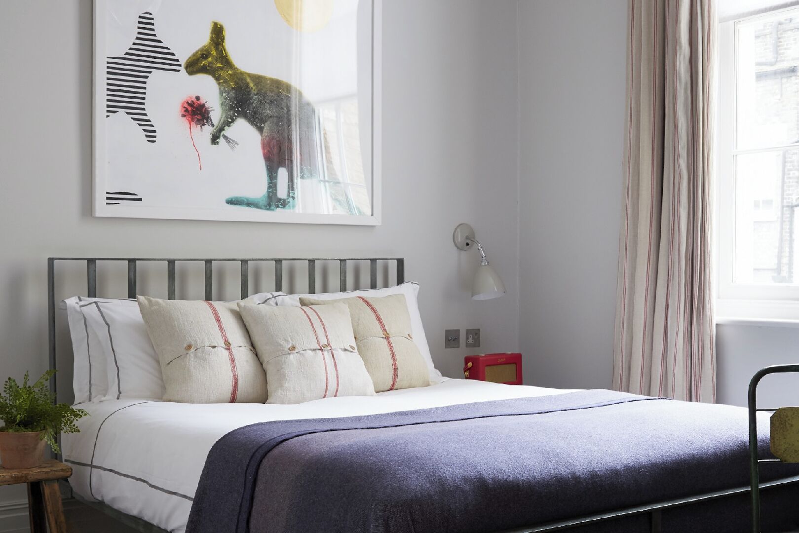 Une chambre à coucher moderne avec un lit, des oreillers, une lampe de chevet, un fauteuil rouge, une plante et un tableau représentant un paysage urbain.