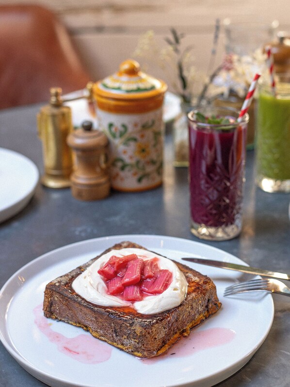 A plate with a slice of toast topped with cream and red fruit, a glass of green smoothie, and a jar of honey. - Artist Residence London (London)