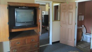 A hotel room with a wooden TV stand, a door leading to a bathroom, and a hallway with a patterned ceiling.