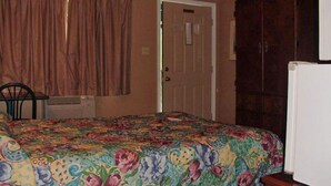 A bedroom with a floral bedspread, a wooden wardrobe, a white refrigerator, and a door with a sign.