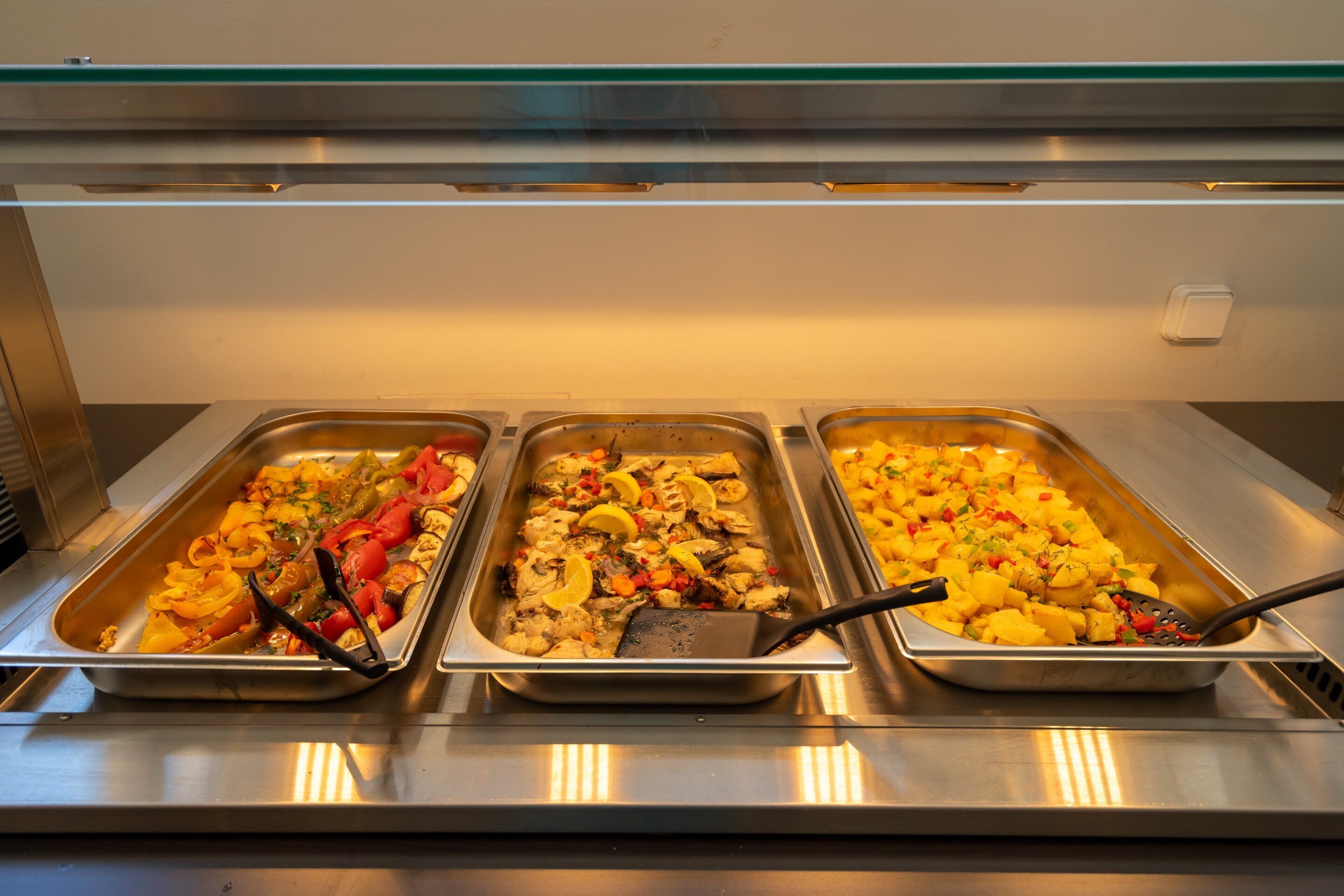A buffet with three trays of food, including vegetables and fish, with serving utensils.