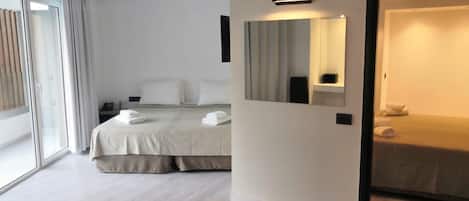 A modern hotel room with a large bed, a bedside table, a mirror, and a view of the outside through a sliding door.