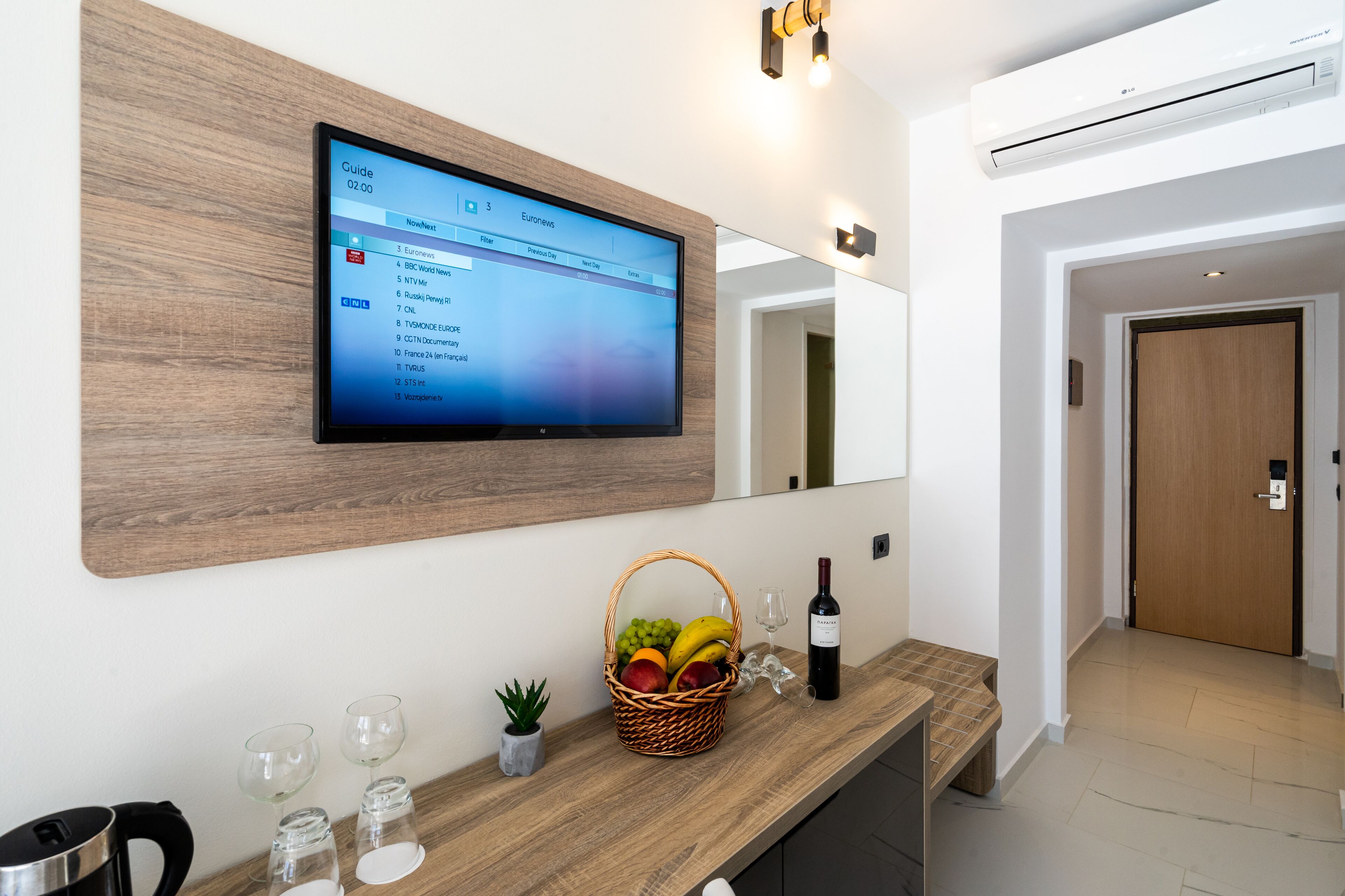 A modern hotel room with a flat-screen TV mounted on a wooden wall, a wooden console table with a wine bottle, glasses, and a fruit basket.