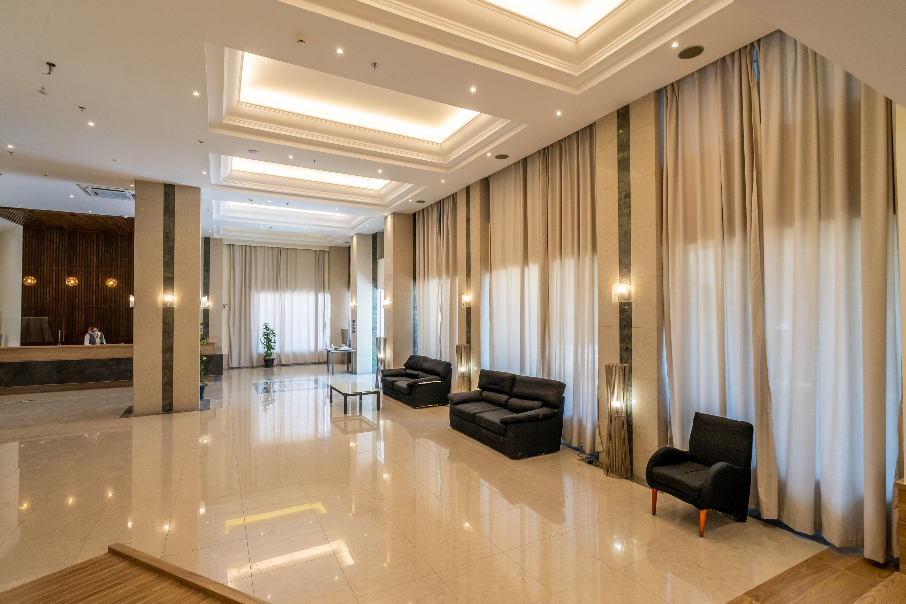 A spacious lobby with a polished floor, two black sofas, a wooden bench, and a reception area with a wooden desk and a plant.