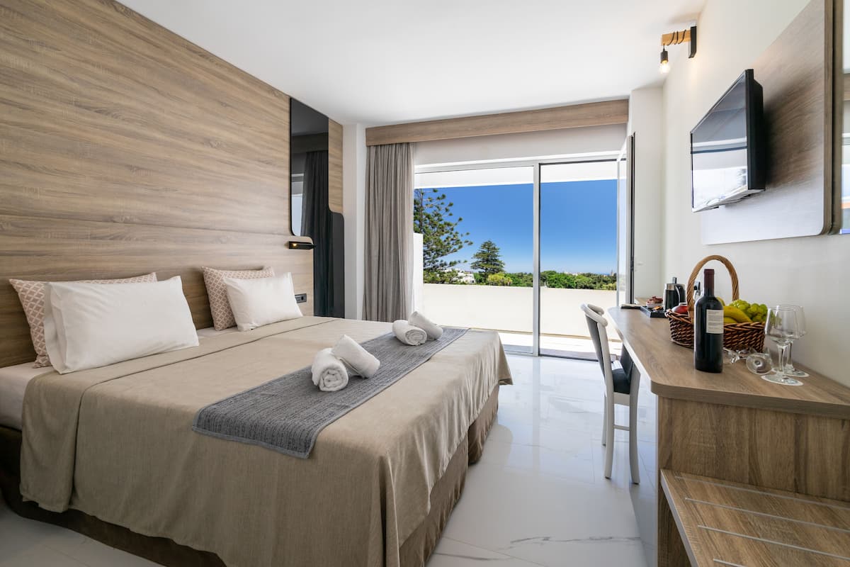 A modern hotel room with a large bed, a desk with a bottle and glasses, and a view of the outdoors.
